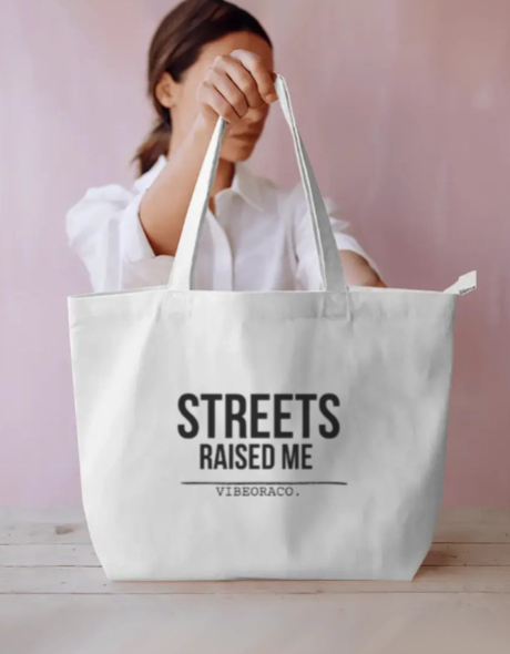 A large off-white canvas tote bag with black text reading 'STREETS RAISED ME' and 'VIBEORACO.' sits on a light wood surface.
